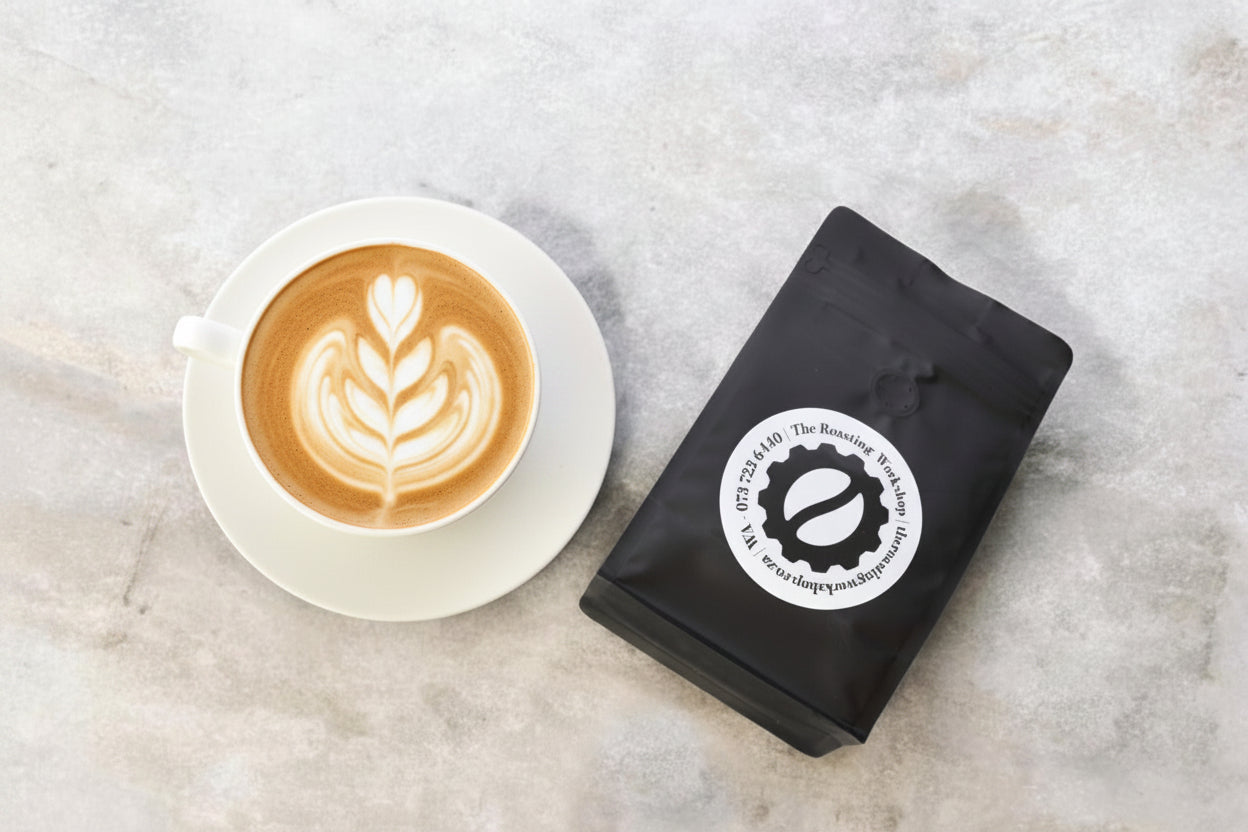 A black coffee bag with a white circular logo sits next to a white cup filled with roasted coffee beans on a light gray stone surface.